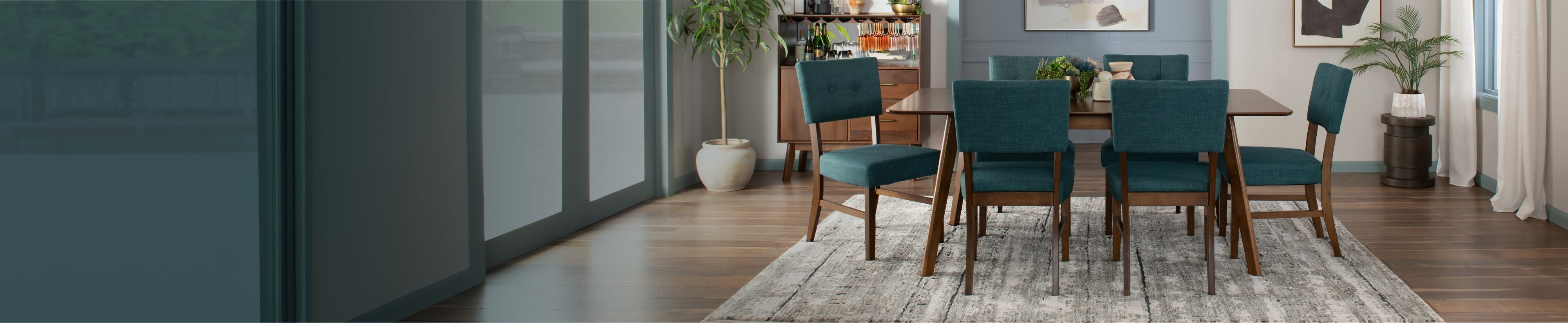 Modern dining room with a wooden table and six teal upholstered chairs on a patterned rug. Art pieces, plants, and natural light create a serene ambiance.
