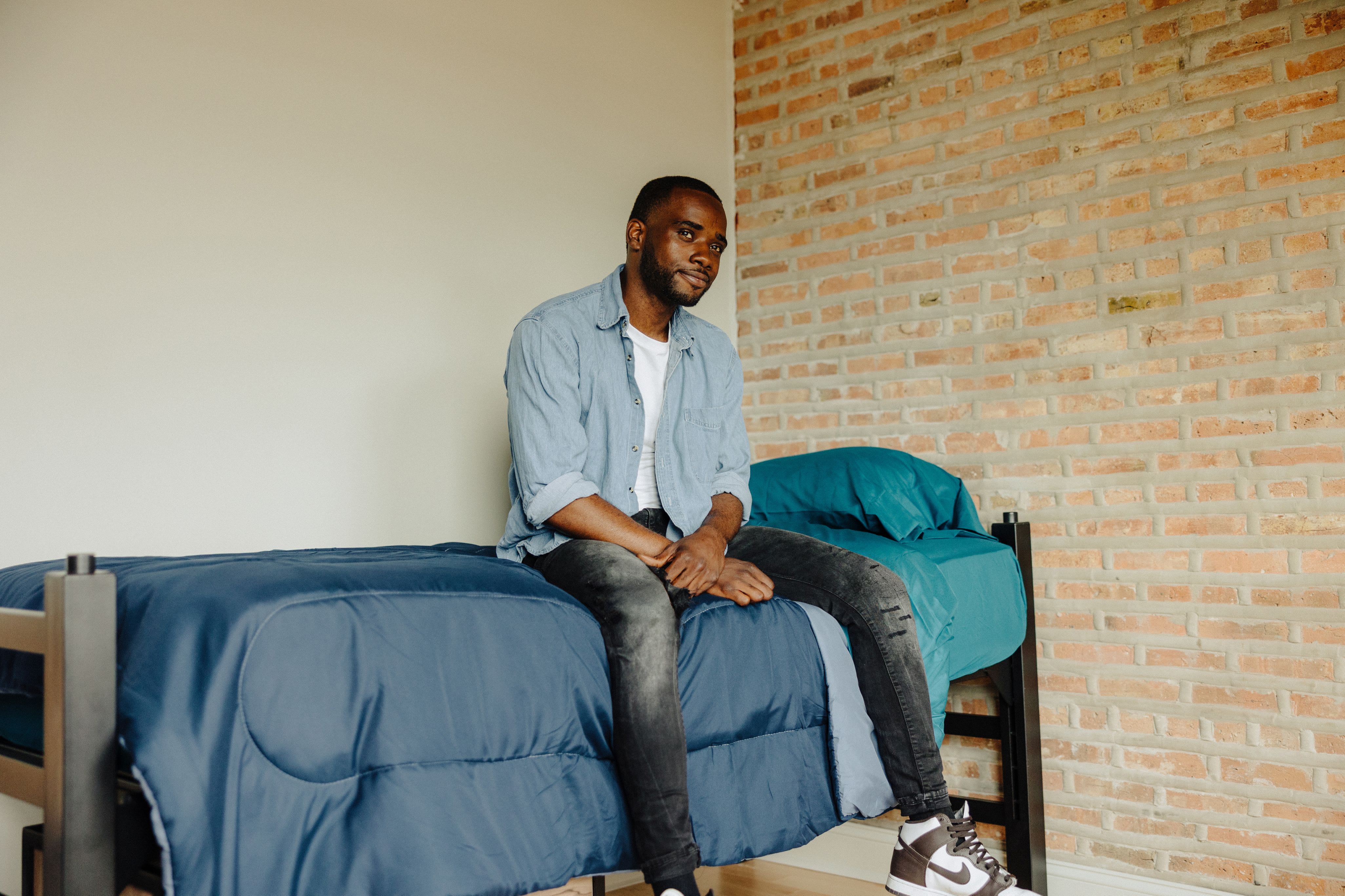 Man wearing a light blue denim shirt, white t-shirt, black jeans, and brown Nike sneakers sitting on a twin bed with blue bedding against a brick wall.