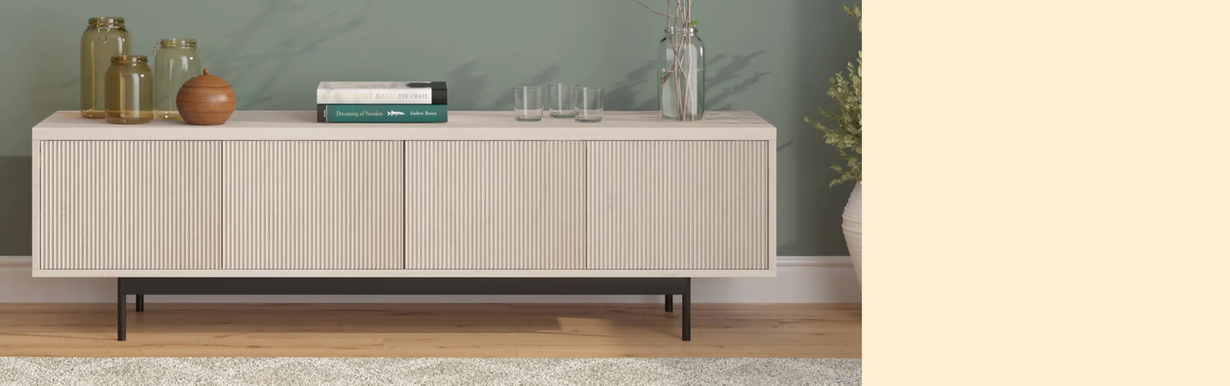Modern living room with a light wood sideboard against a sage green wall. Decor includes vases, books, and glasses. Cozy, minimalist style.