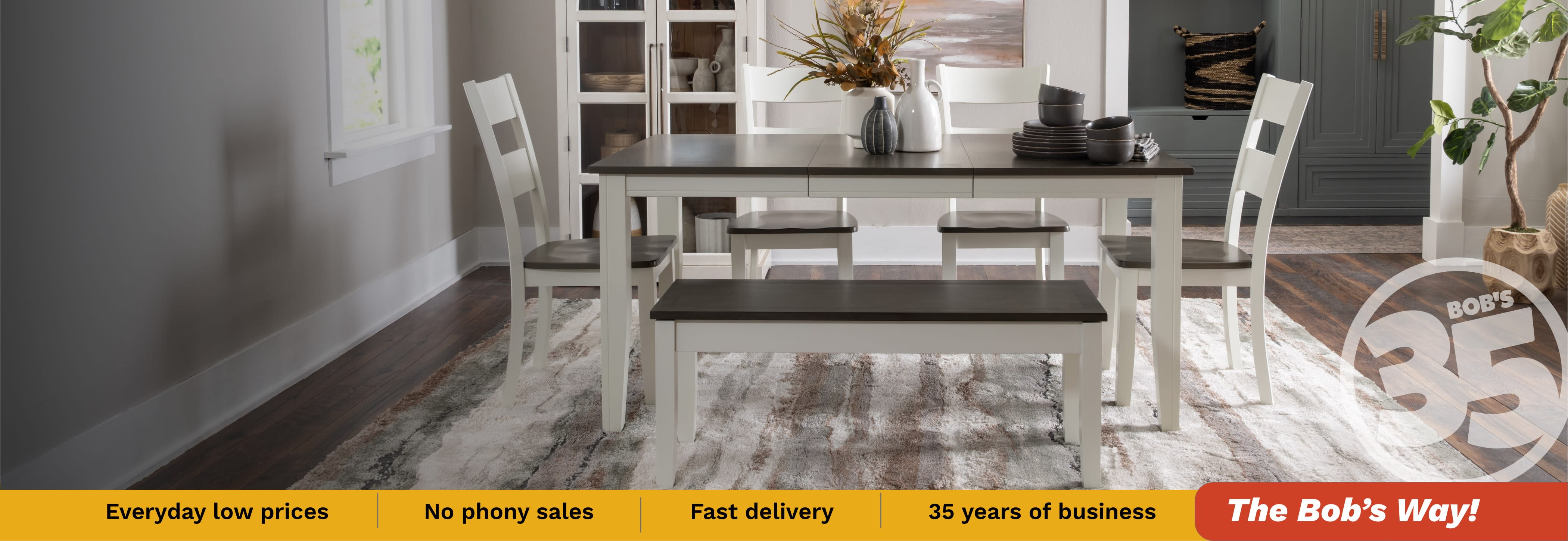 Bright modern dining room with a white table, benches, chairs, floral centerpiece, and dinnerware on a patterned rug; calm, inviting decor.