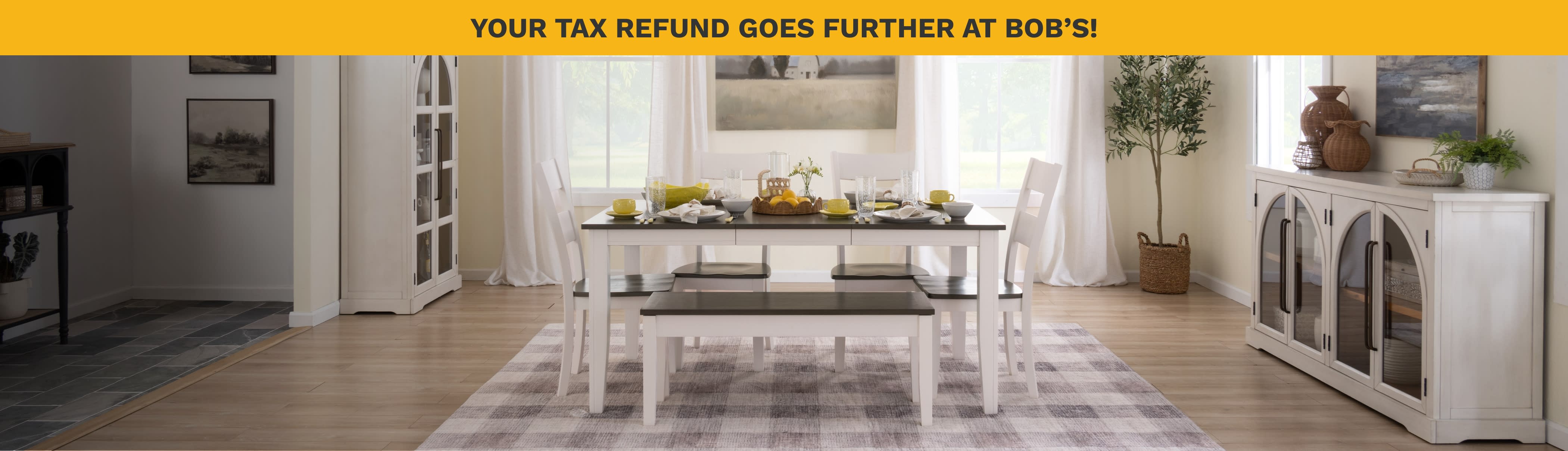 A cozy dining room with a white table, chairs, and bench on a checkered rug. A centerpiece of bread and lemons adds warmth. Two large windows flank a farmhouse painting, creating an inviting, serene atmosphere.