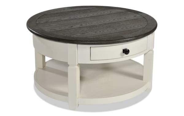 Laurel White & Brown Round Storage Coffee Table from Laurel Entertainment & Occasional collection, farmhouse style MDF wood with two-tone finish, distressed dark brown top, white base, black hardware drawer, and lower shelf for living room storage.