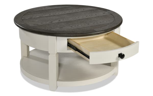 Laurel White & Brown Round Storage Coffee Table from Laurel Entertainment & Occasional collection, farmhouse style MDF wood with two-tone finish, distressed dark brown top, light base, open shelf, and a small storage drawer with black hardware.
