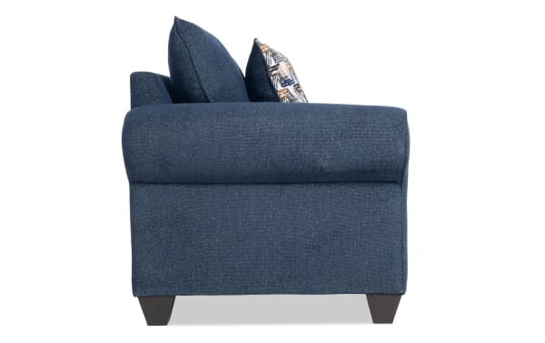 Side view of the Kylie Navy Chair in transitional style, featuring navy blue polyester fabric upholstery, rolled arms, tapered dark legs, and plush accent pillows.