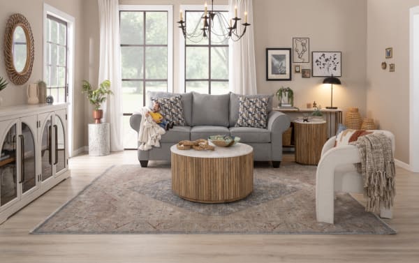 Living room with a gray fabric polyester sofa featuring patterned cushions, a round wooden coffee table, light wood flooring, and neutral decor in transitional style.