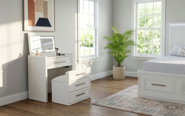 White wooden vanity desk with cushioned stool, large mirror, and makeup items next to a bed with storage drawers, hardwood floor, and potted plant in a bright bedroom.