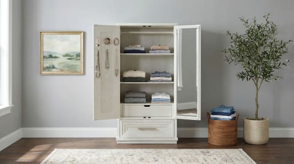 Light wood wardrobe with open doors showing neatly folded sweaters and shirts on shelves, a drawer, and a full-length mirror on one door, set against a gray wall with a framed landscape painting, a woven basket with folded clothes, and a potted green plant on dark hardwood flooring.