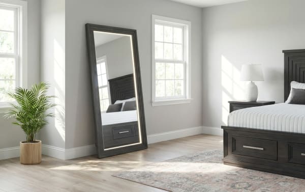 Modern bedroom corner with a large dark wood framed LED lighted standing mirror, light gray walls, hardwood floors, potted green plant, and a dark wood bed with white bedding.