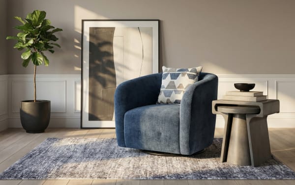 Modern living room corner with a plush navy blue velvet swivel armchair featuring a geometric patterned cushion, next to a concrete side table with stacked books and a black bowl, set on a textured gray area rug. Includes a large abstract framed artwork leaning against the wall and a potted fiddle leaf fig plant on light wood flooring.