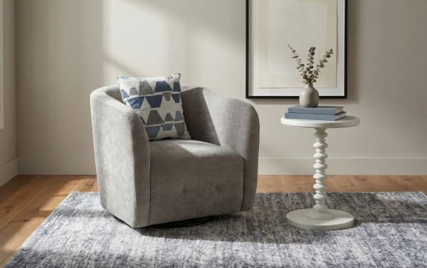 Gray upholstered swivel armchair with a blue and white geometric patterned cushion, next to a white round pedestal side table with books and a vase, on a textured gray rug over wood flooring.