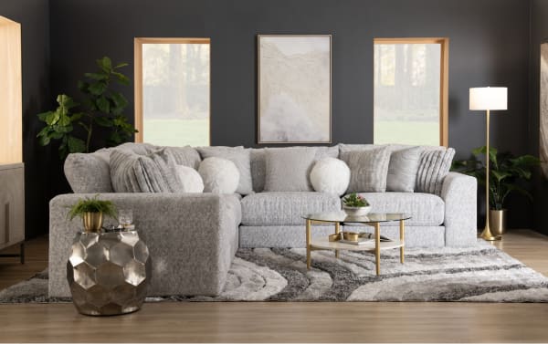 Gray sectional sofa with textured fabric and multiple decorative pillows, paired with a round glass coffee table and metallic side table on a patterned gray rug in a modern living room.