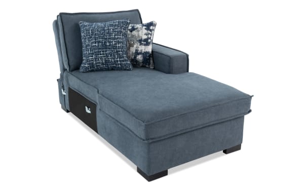 Blue upholstered chaise lounge with two decorative patterned throw pillows, featuring a cushioned armrest and storage compartments underneath.