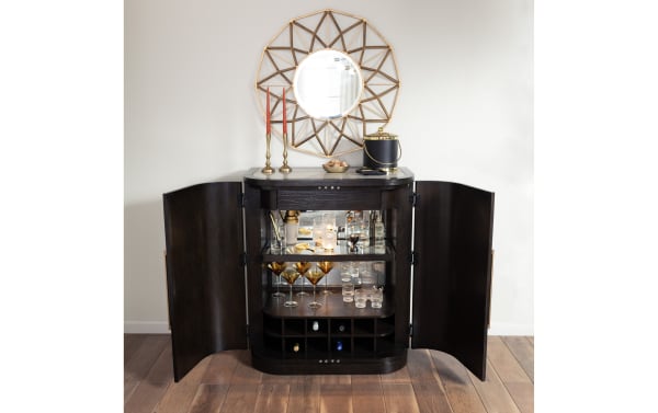 Dark wood bar cabinet with open doors, featuring glass shelves with cocktail glasses, bottles, and gold accents, under a geometric gold-framed mirror.