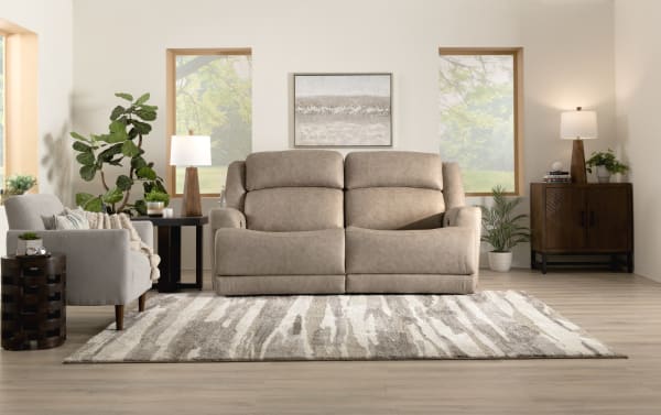 Modern living room setup with a beige leather reclining loveseat, gray fabric armchair, wooden side tables, patterned area rug, and natural light from three windows.