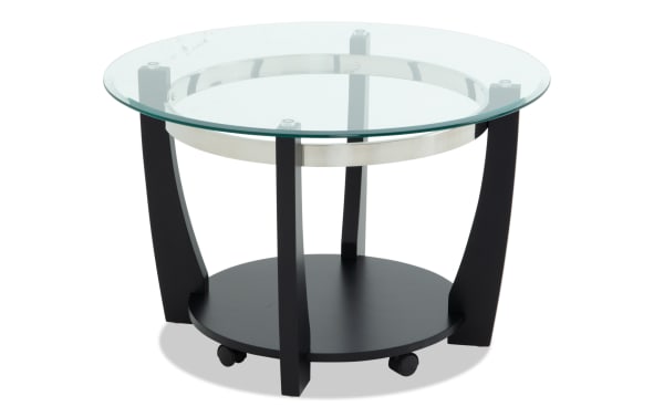 Round modern side table with a clear glass top, black wooden legs, a lower black shelf, and caster wheels for mobility.