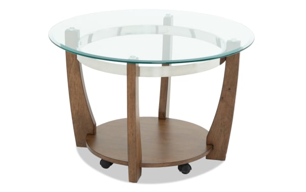 Round wooden coffee table with a glass top and lower wooden shelf, featuring curved legs and caster wheels for mobility.