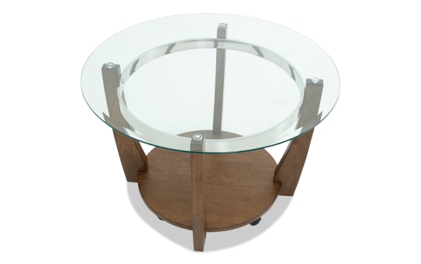 Round glass-top side table with wooden legs and lower wooden shelf, featuring a modern design with metal accents on the glass supports.