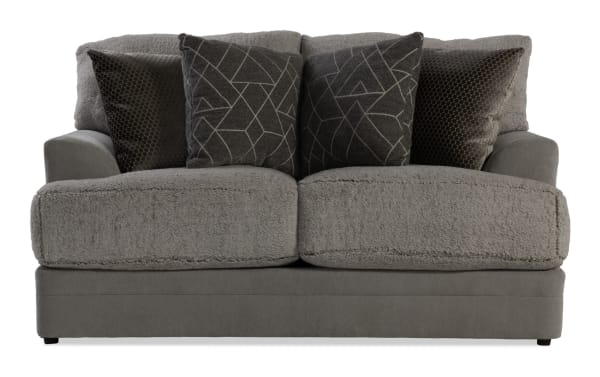 Gray plush fabric loveseat with thick cushioned seats and backrests, featuring four decorative throw pillows in geometric and textured patterns.