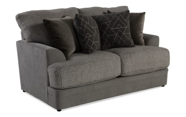 Gray plush loveseat sofa with soft textured cushions and four decorative throw pillows in dark geometric and honeycomb patterns, modern style.