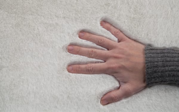 Hand pressing down on plush white Angora 5' x 7' solid transitional rug with high-pile polyester fabric and cushioned memory foam backing for comfort and odor control.