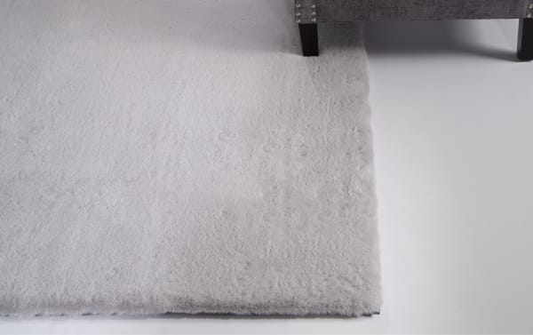 Soft, plush light gray area rug with a thick pile, placed on a smooth white floor next to a dark gray upholstered chair leg.