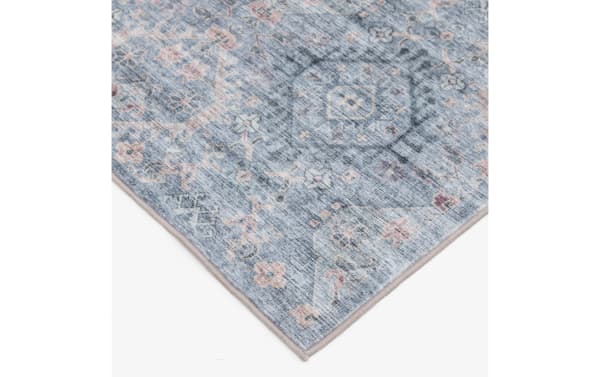Corner of a blue and gray vintage-style area rug with intricate floral and geometric patterns in muted pink and beige tones, featuring a soft woven texture and neatly finished edges.