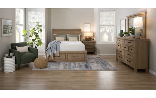 Bedroom with light wood furniture including a bed with storage drawers, matching nightstand and dresser with mirror, green armchair, and decorative plants.
