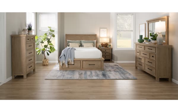 Modern bedroom set with light wood furniture including a bed with storage drawers, matching nightstand, dresser with mirror, chest of drawers, and decorative plants on a light wood floor.