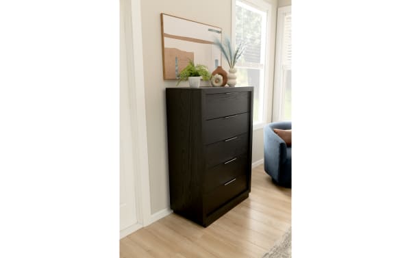 Black wooden chest of drawers with four metal handles, decorated with a potted fern and modern vases, placed in a bright room with light wood flooring and large windows.