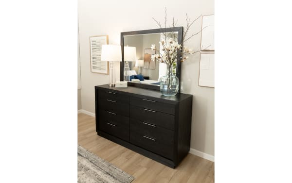 Dark wood dresser with eight drawers and sleek metal handles, topped with a large framed mirror, glass lamp, and vase with white flowers, in a modern bedroom setting.