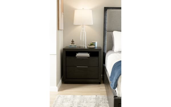 Dark wood nightstand with two drawers and metal handles, topped with a clear glass table lamp, small plant, and decorative items, next to a gray upholstered bed.
