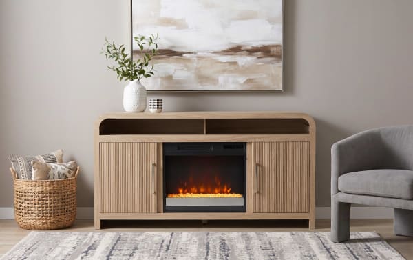 Modern wooden media console with built-in electric fireplace, fluted cabinet doors, light oak finish, decorative vase, and gray armchair in living room setting