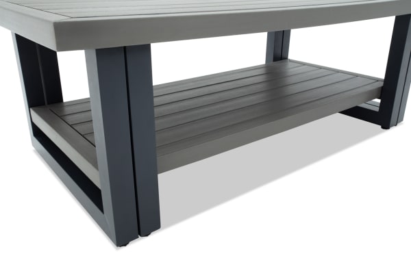 Modern outdoor coffee table with a gray concrete top, black metal frame legs, and a lower gray wooden slatted shelf for storage.