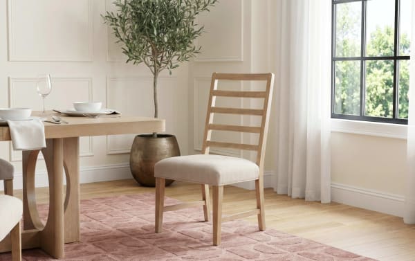 Wooden dining chair with beige upholstered seat and ladder-back design, placed on a textured pink rug next to a light wood dining table and window.