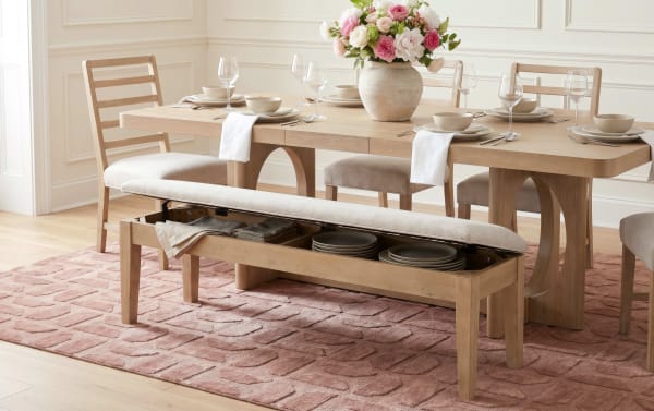 Light wood dining table set with beige cushioned chairs and a storage bench featuring a lift-up seat holding stacked plates and linens, placed on a textured pink rug. The table is set with beige dishware, wine glasses, silver cutlery, white napkins, and a large floral centerpiece with pink and white flowers.