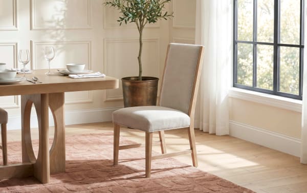 Dining chair with beige upholstered seat and backrest, wooden frame, placed next to a light wood dining table on a textured brown rug in a bright room with large window and potted plant.