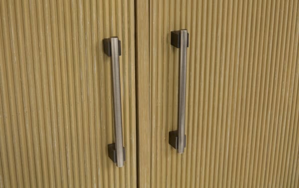 Vertical brushed metal cabinet handles mounted on textured light brown wooden cabinet doors with vertical grooves.