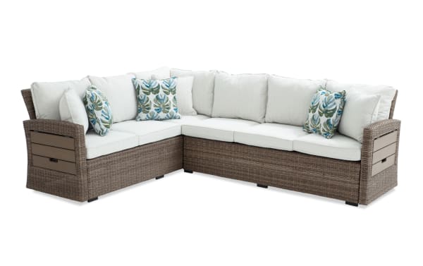 Outdoor sectional sofa with brown wicker frame, beige cushions, and tropical leaf-patterned throw pillows, designed for patio seating.