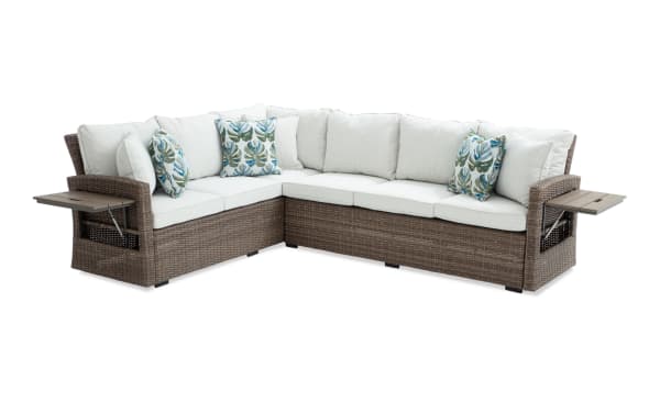 Outdoor sectional sofa with brown wicker frame, white cushions, and tropical leaf-patterned throw pillows, featuring built-in side tables for patio use.