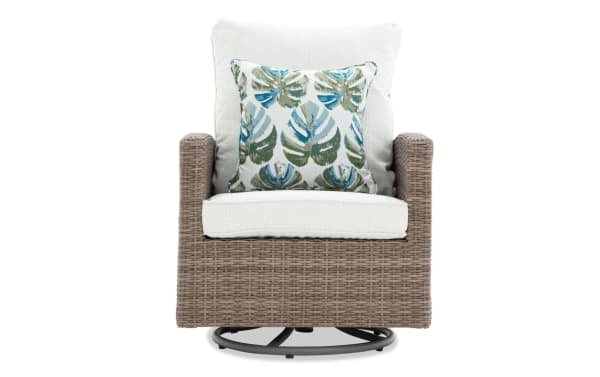 Wicker swivel chair with beige cushions and a decorative pillow featuring green and blue tropical leaf patterns, ideal for outdoor or patio seating.