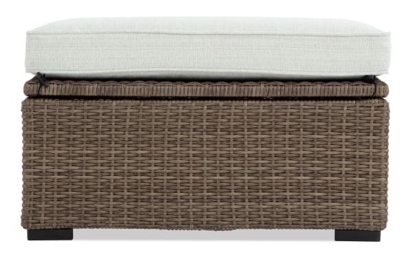 Outdoor wicker storage ottoman with light beige cushioned top, brown woven rattan base, and black feet, suitable for patio seating and storage.