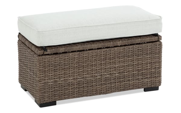 Outdoor storage ottoman with woven rattan wicker base in brown and removable beige cushion, ideal for patio seating and storage.