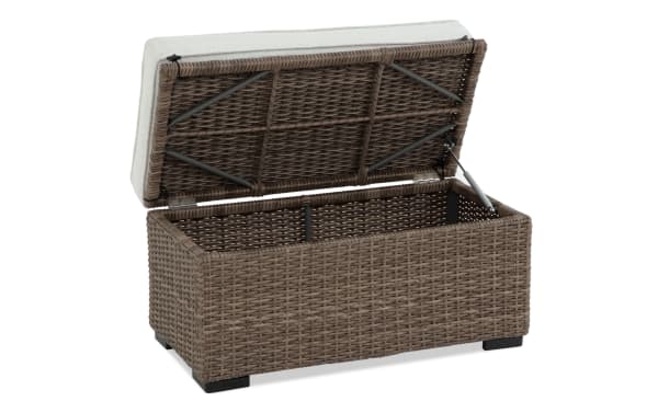 Outdoor storage bench with brown woven rattan wicker, beige cushioned lid, metal frame support, and black feet, ideal for patio organization.