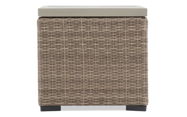 Outdoor side table with light gray wooden top and woven synthetic rattan base in taupe color, featuring black feet for patio or garden use.