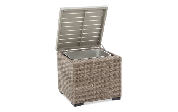 Wicker outdoor storage ottoman with hinged lid and inner metal cooler tray, featuring a beige woven rattan design and black feet.
