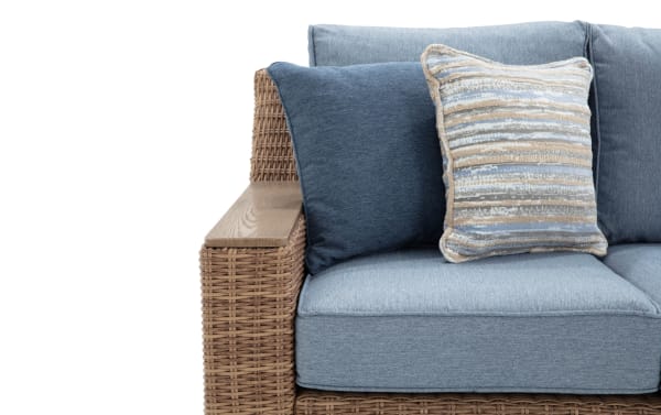 Close-up of a blue-gray fabric outdoor sofa with woven rattan armrest and two decorative cushions, one striped in neutral tones.