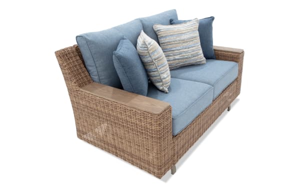 Blue cushioned two-seater wicker sofa with wooden armrests, featuring multiple decorative pillows in blue and beige tones, ideal for outdoor or patio seating.