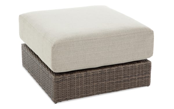Beige Modular Bob® outdoor ottoman with brown all-weather wicker base, rust-proof aluminum frame, and plush polyester cushion.