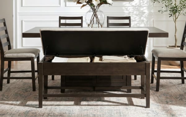 Dark wood storage bench with cushioned seat and hinged top, shown open with folded linens inside, placed in a dining room with matching chairs and a wooden table.