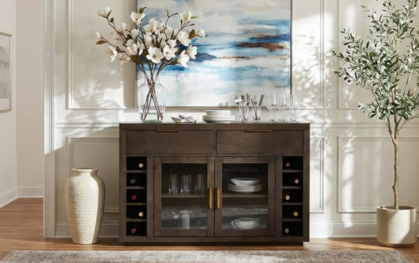 Dark wood sideboard with glass doors and brass handles, featuring built-in wine racks, white plates, glassware, and decorative vases with flowers, set against a white paneled wall with a blue abstract painting.